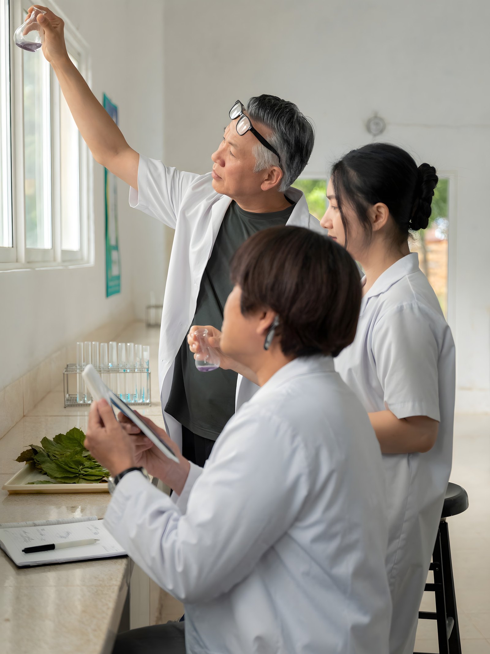 Three researchers in white coats in a bright laboratory: one holds a small flask up to the window light, one watches, one takes notes
