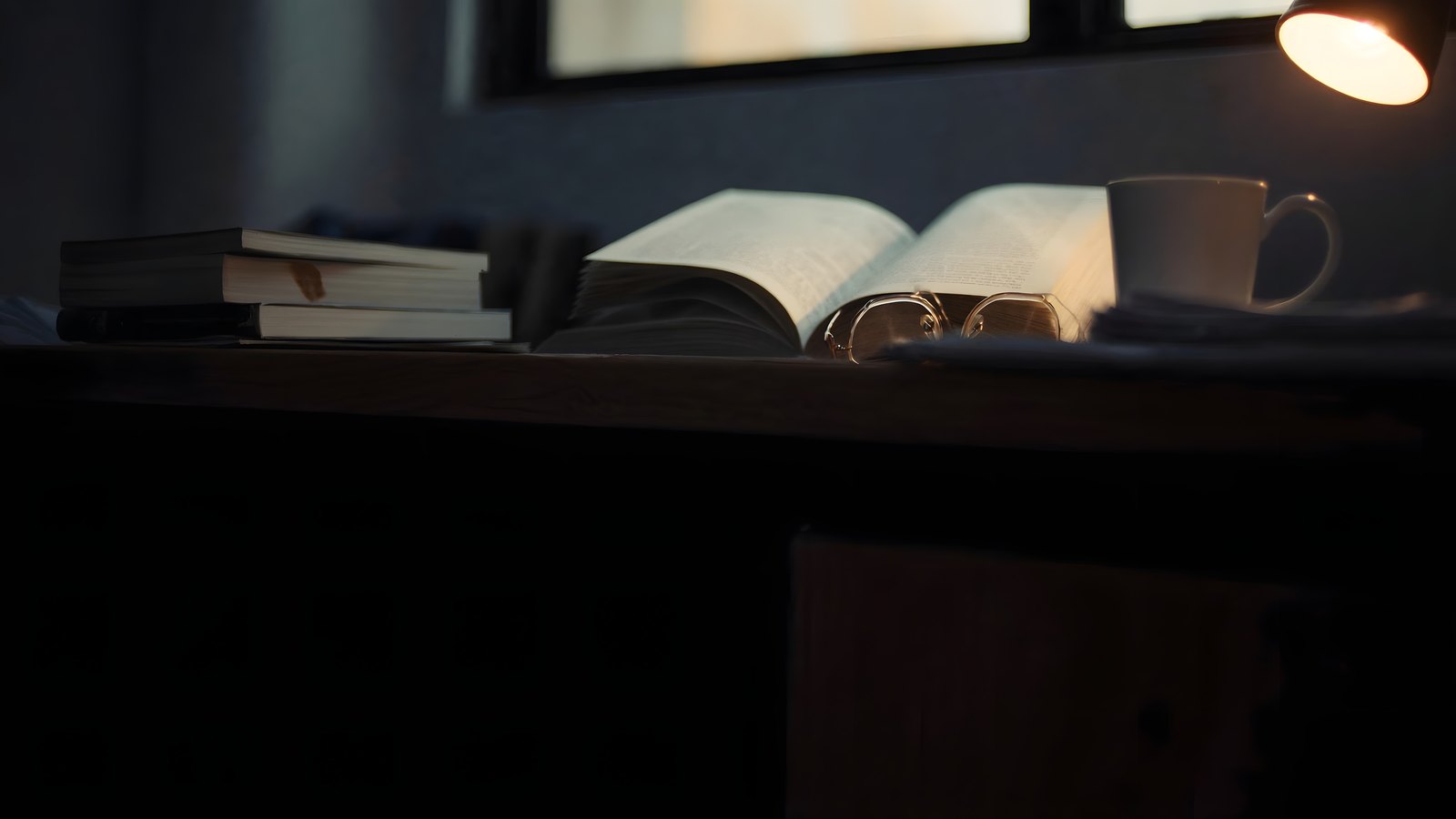 An open journal, reading glasses and a cup on a desk by a window at dusk, under a small lamp
