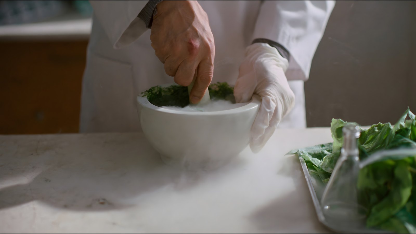 Gloved hands grind frozen green leaf in a white mortar as cold vapour spills across the bench