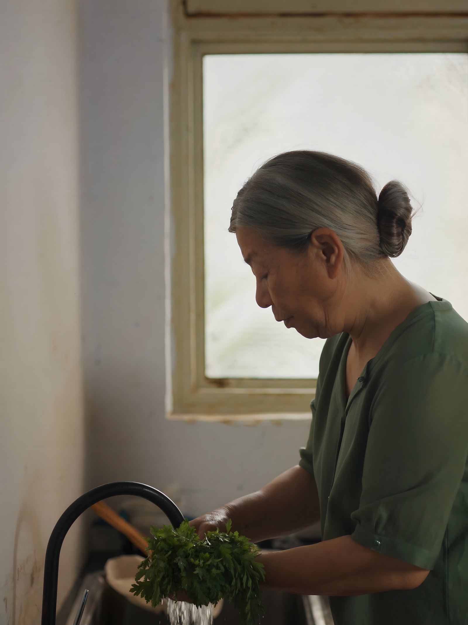 A grey-haired woman in a green blouse rinses a bunch of herbs under the tap at her kitchen sink, by a window