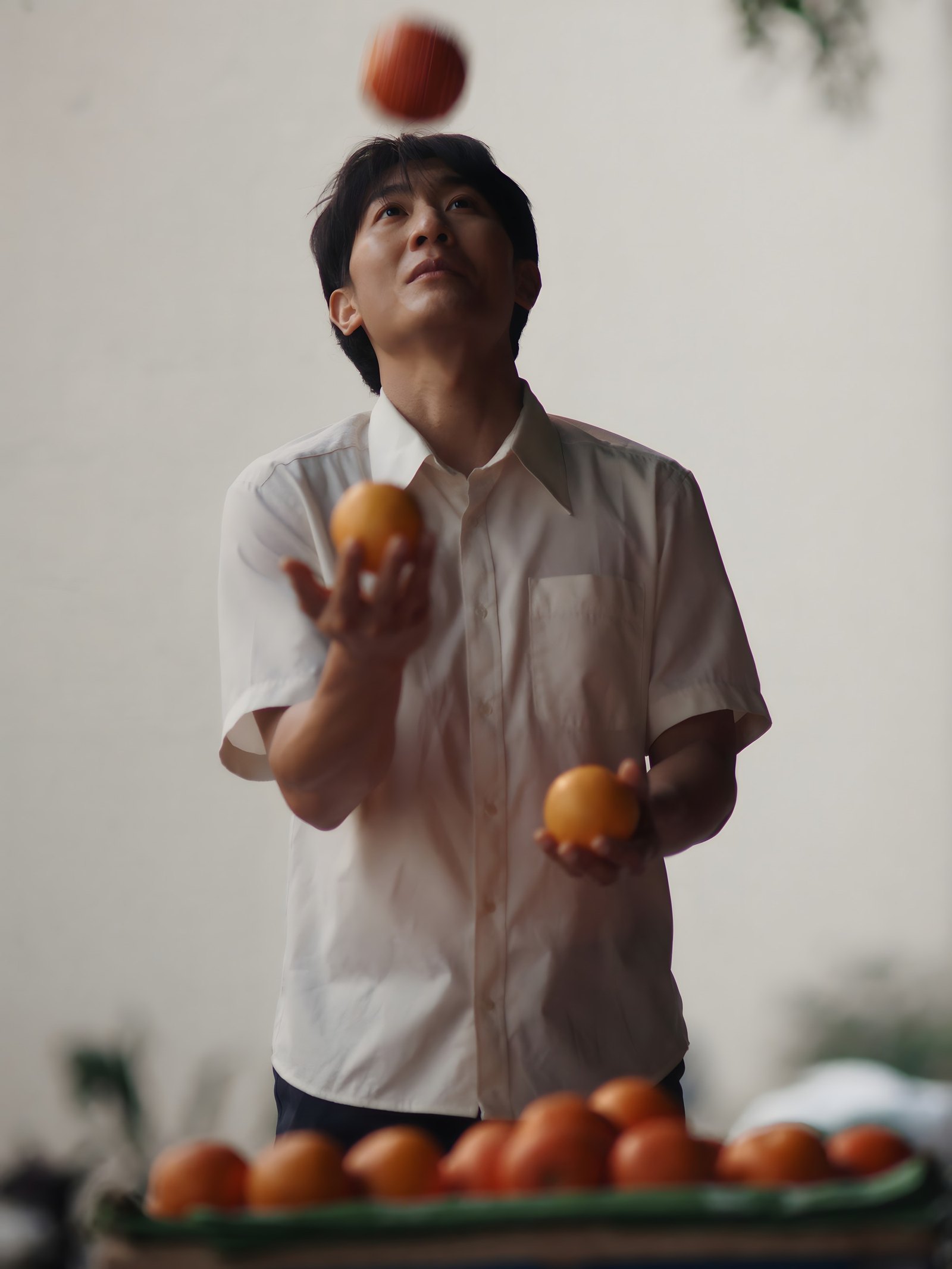 A young man in a white shirt juggles oranges above a tray of oranges, his eyes on the highest one